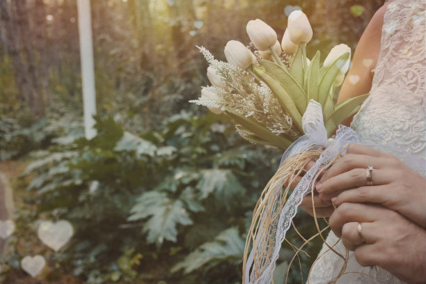 新娘和新郎手持优雅的现代秋季婚礼花束前面的白色婚纱。背景中可以看到新娘和新郎的结婚戒指图片下载