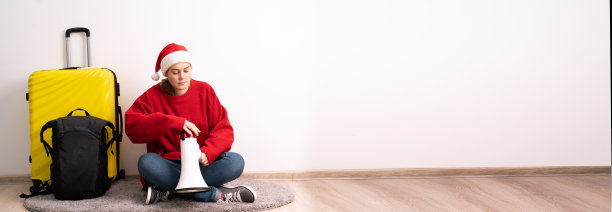带着圣诞老人帽子的行李箱的女孩旅行者坐在地板上放着扬声器。圣诞节概念门票和旅游销售。横幅图片下载