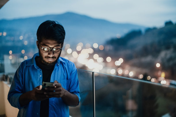 一名戴眼镜、穿蓝色衬衫的印度男子晚上在一座现代建筑的阳台上使用智能手机。在城市夜街的背景中图片下载