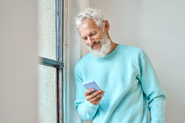 微笑的老胡子潮人使用手机站在窗口。图片下载