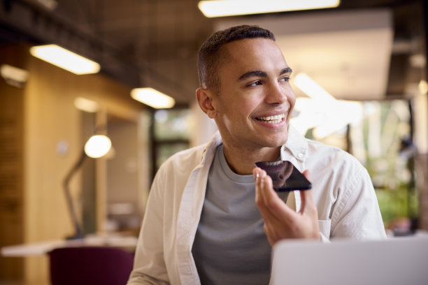 年轻的商人在办公桌上的笔记本电脑上工作，对着手机的麦克风说话图片下载