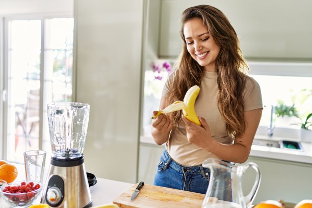 年轻美丽的西班牙女子准备蔬菜奶昔与搅拌机剥香蕉在厨房图片下载