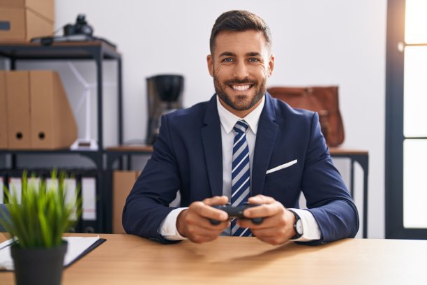 英俊的西班牙人在办公室玩电子游戏，脸上带着快乐和凉爽的微笑。显示的牙齿。图片下载