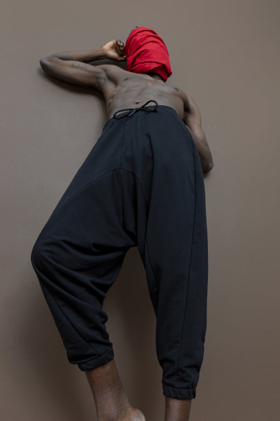 黑人赤裸上身站在棕色的墙前，用红布盖住他的头。图片下载