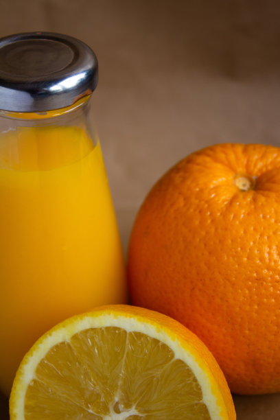 一瓶橙汁有一个闪亮的盖子，水果本身和半个橙子在浅棕色模糊的背景-垂直照片图片下载
