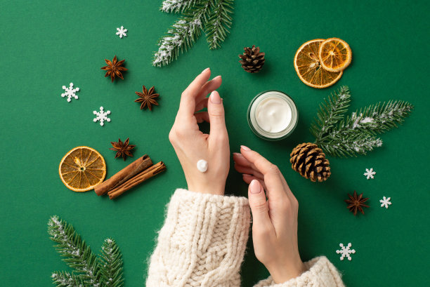 第一人称俯视图照片的女孩在毛衣使用护手霜从小罐子松果干橘子片肉桂棒八角雪花和云杉树枝在雪孤立的绿色背景图片下载