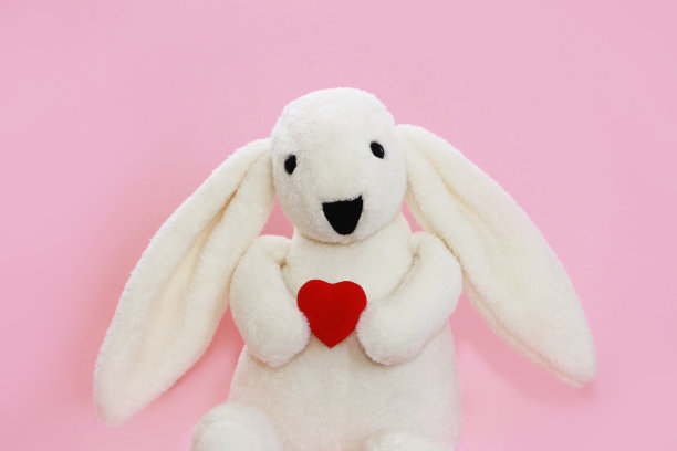 儿童玩具柔软蓬松的白兔抱着粉红色背景上的红心图片下载