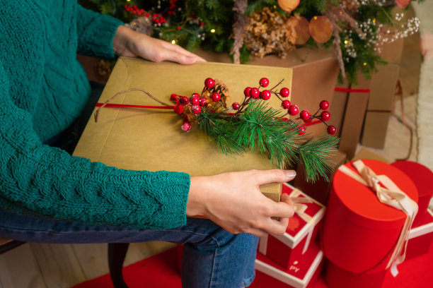 一名身穿绿色毛衣和蓝色牛仔裤的女子抱着一个手工包装的礼物，她的腿上放着一棵圣诞树，圣诞树上有红色的礼品盒。圣诞节和新年的概念图片下载