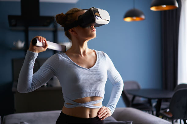 年轻的未来金发女孩戴着虚拟现实耳机，拿着控制器在家里玩视频游戏。女子在客厅中间玩虚拟现实虚拟现实射击战斗。图片下载