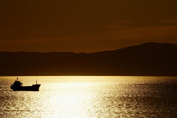 一个明亮的暗青铜日落在海上，上面有工业船和帆船。海参崴海上的日落。海上的日落进入了群山。图片下载