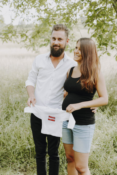 一名孕妇和她的男朋友站在绿色草地上，手里拿着白色的婴儿衬衫，上面有奥地利国旗图片下载