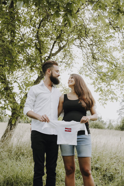 一名孕妇和她的男朋友站在绿色草地上，手里拿着白色的婴儿衬衫，上面有奥地利国旗图片下载