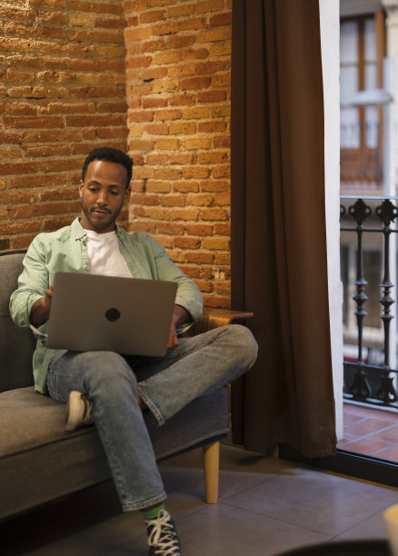 英俊的黑人非裔美国人坐在舒适的客厅沙发上，在笔记本电脑上工作。自由职业者在家工作，使用社交网络。-垂直照片-图片下载