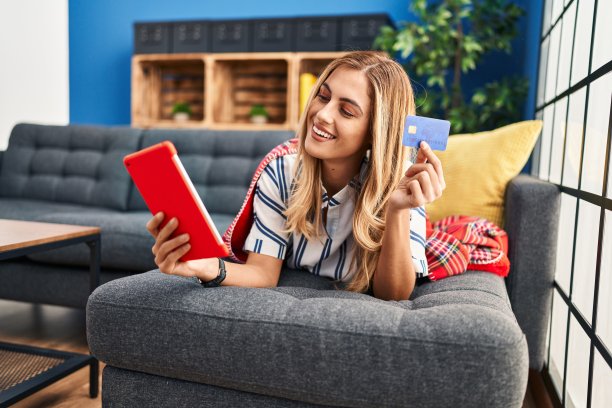 年轻的金发女子使用笔记本电脑和信用卡坐在家里的沙发上图片下载