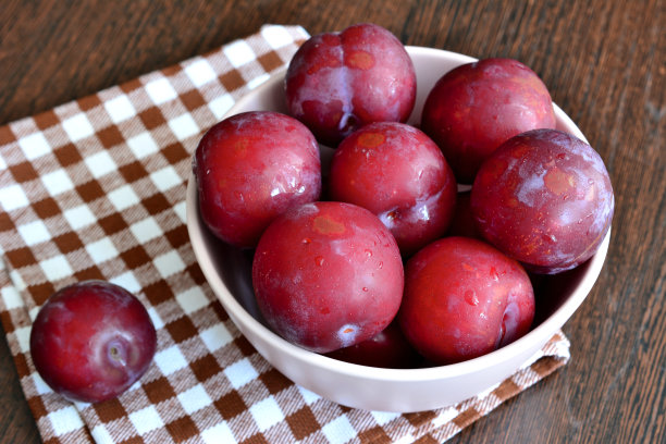 紫色李子放在碗里，厨房餐巾在深色的木制背景上图片下载