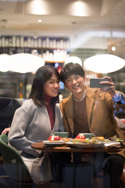 冬天，年轻的东亚夫妇坐在一家餐厅里愉快地聊天，用智能手机自拍——库存照片图片下载