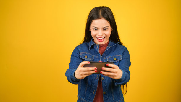 快乐的现代女孩使用手机的肖像，年轻女子在智能手机上玩视频游戏，孤立地站在黄色背景上图片下载