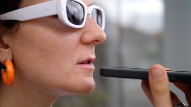 一名年轻女子戴着时髦的白色太阳镜和橙色耳环，她的面部特写在语音记录仪上录制语音信息，以便进一步发送给她的对话者。图片下载