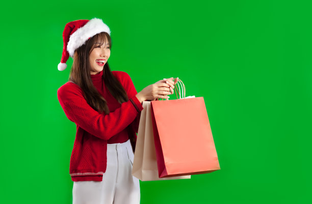 年轻的亚洲妇女在红白相间的衣服，戴着圣诞老人帽子，手持和携带纸质购物袋在绿色屏幕背景。圣诞快乐主题。图片下载