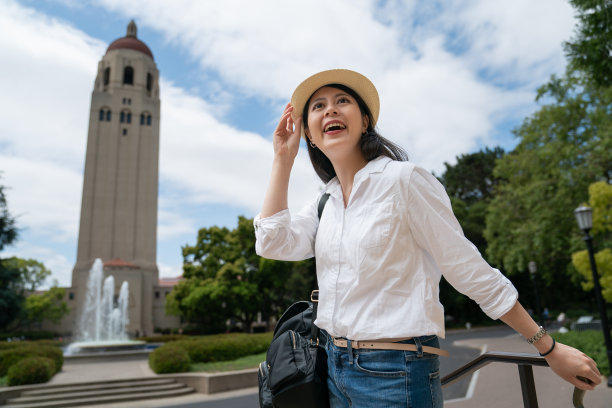 cheerful asian woman traveler holding hat and looking up at sunny sky with hoover fountain and bell tower at background. sheâs happy with the nice weather during her visit on school campus图片下载