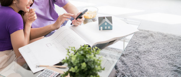 年轻幸福的恩爱夫妇一边聊天，一边用平板电脑计算买房的价格，房子图纸上有一个小木屋模型。新家居概念。图片下载