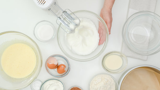 将搅拌好的蛋白放入玻璃碗中。一步步制作巧克力蛋糕的配方，烘焙过程。白色餐桌背景图片下载