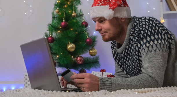一名男子用笔记本电脑和信用卡为新年和圣诞节购买礼物。一名戴着圣诞帽的男子站在圣诞树的背景上，手里拿着笔记本电脑，还有花环和礼物。来自骗子的网络安全。图片下载