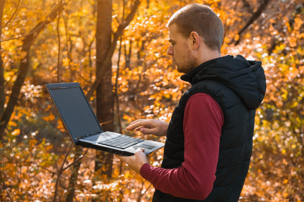 一个自由职业者在秋天的森林里远程工作。乡村生活。脱离文明。范Lifevibes。一个拿着笔记本电脑的男人。通过笔记本电脑在新鲜空气中进行远程学习。远程工作。图片下载