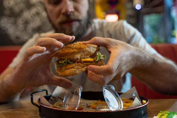 留着胡子的白人中年男子，在咖啡馆或餐厅吃汉堡和快餐。汉堡不是有益的食物。很饿的家伙。饮食的概念。不健康食品的概念，小吃，街头小吃。图片下载