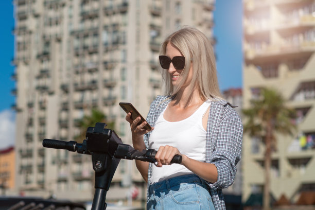 年轻微笑的金发女子戴着太阳镜在一个阳光明媚的日子里用手机打开一辆电动滑板车，复制空间图片下载