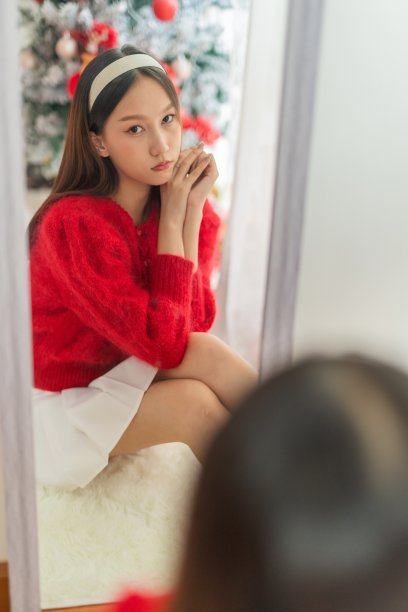 年轻美丽友好的亚洲女性女士，红色长袖毛衣和白色裙子，漂亮的发带，在镜子前摆出不同的样子，在她的房间里有一棵装饰精美的圣诞树作为背景图片下载