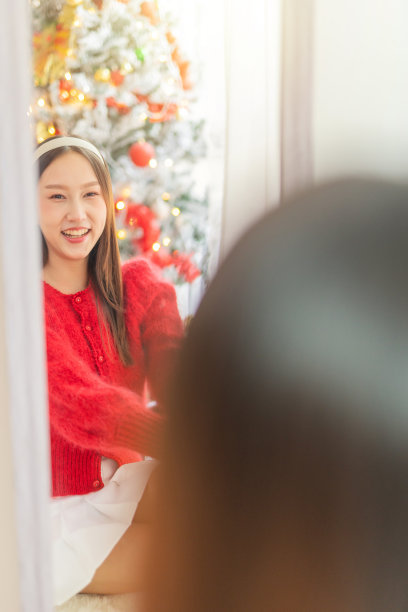 年轻美丽友好的亚洲女性女士，红色长袖毛衣和白色裙子，漂亮的发带，在镜子前摆出不同的样子，在她的房间里有一棵装饰精美的圣诞树作为背景图片下载