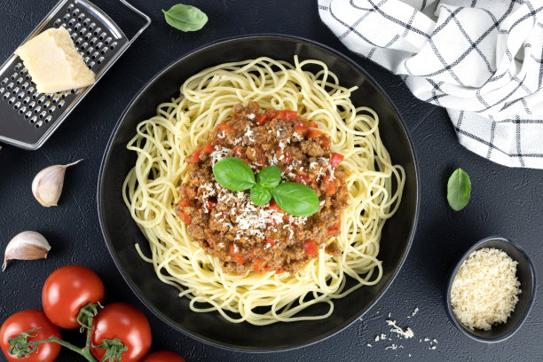 意大利肉酱面放在黑色盘子里，黑色背景上有新鲜的罗勒、西红柿、大蒜和帕玛森奶酪。图片下载