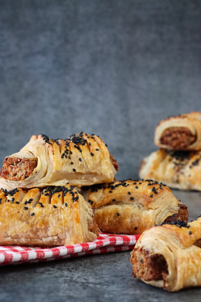 近距离图片，一批烘烤的自制香肠卷，上面撒着黑芝麻，在红白格子茶巾上，斑驳，灰色背景，重点在前景图片下载
