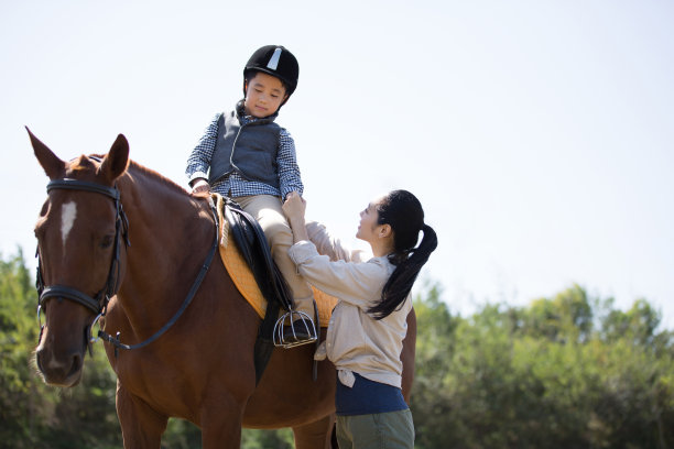 在秋高气爽的天空下，母亲和儿子在草地上练习骑马，男孩骑在马背上，母亲深情地教导-资料照片图片下载