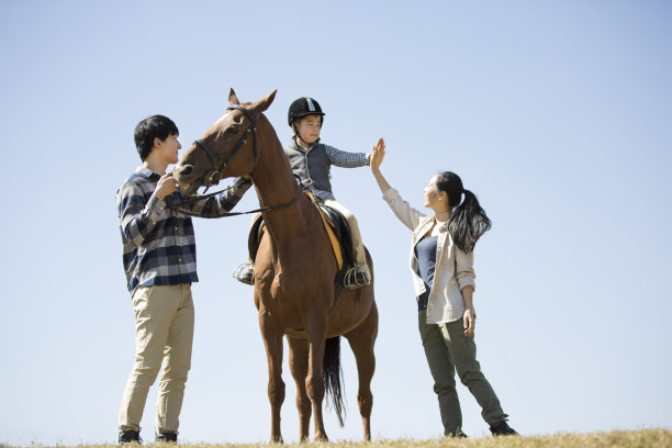 在晴朗的天空下，年轻的家庭在马场练习马术和骑马，儿子和母亲击掌庆祝-资料照片图片下载