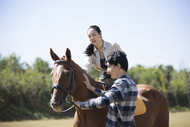 秋高气爽的天空下，一对年轻夫妇在草地上，女士在练习骑马，男子牵着马图片下载