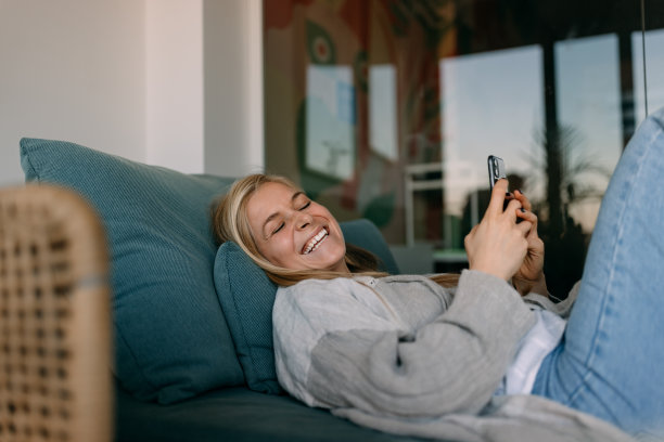 金发女性笑着拿着电话，躺在沙发上。图片下载