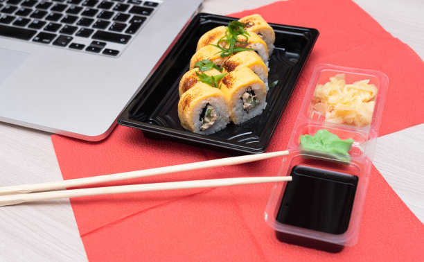 美味的日式卷，里面有三文鱼、牛油果、黄瓜、奶酪，外带包装，还有木棍、酱油和笔记本电脑。有选择性的重点。亚洲外卖食品概念图片下载