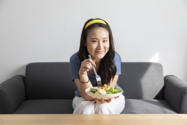 美丽的微笑的亚洲女人手里拿着一盘沙拉，坐在沙发上。图片下载