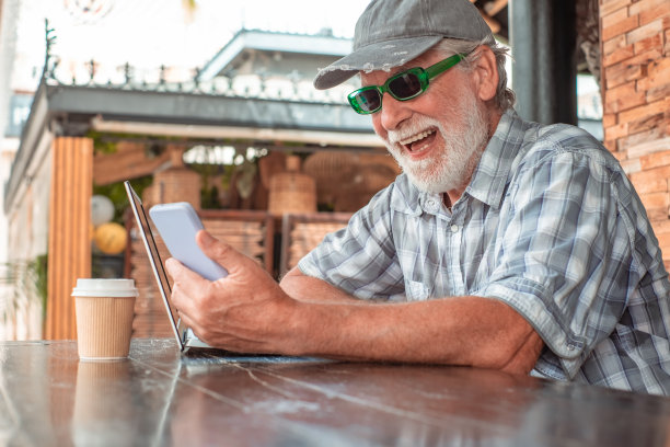 一位开朗的老人坐在一张木桌边，一边用手机谈笑风生。帅气的老男人一边喝咖啡一边在笔记本电脑上上网图片下载