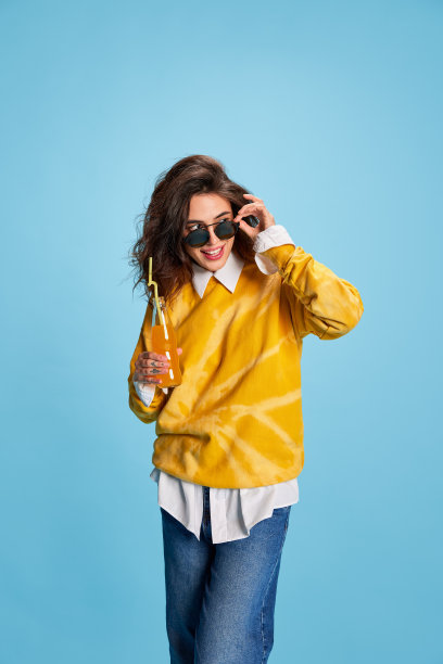 年轻美丽的女孩的肖像在太阳镜和橙汁孤立的蓝色背景。好心情图片下载