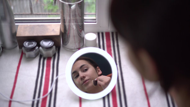 一位年轻女子站在窗前，看着一面小镜子，用刷子化妆。从镜子里看到的女孩。图片下载