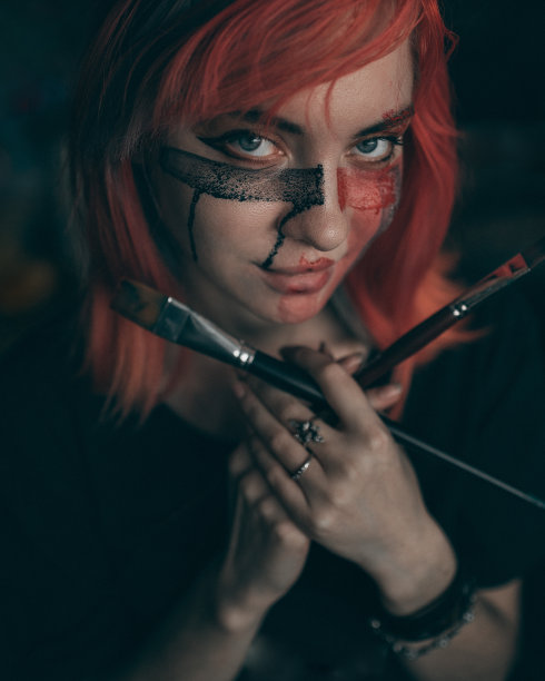 年轻的红发女艺术家用刷子在脸上涂上红色和黑色的颜料图片下载