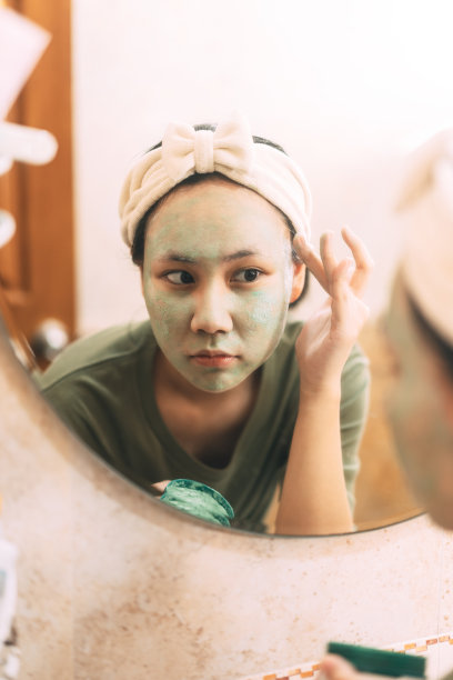 年轻的成年亚洲妇女洁面与绿色粘土面膜愈合痤疮反射与镜子在浴室图片下载
