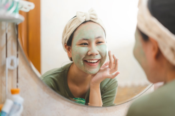 年轻的成年亚洲妇女洁面与绿色粘土面膜愈合痤疮反射与镜子在浴室图片下载