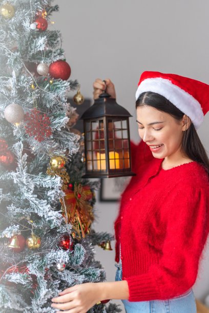 肖像照片的年轻美丽友好的亚洲女性女士与圣诞老人帽手持户外灯笼烛光旁边一个漂亮的装饰圣诞树图片下载