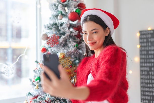 一位年轻美丽友好的亚洲女性女士的肖像照片，戴着可爱的圣诞老人帽，在她房间里装饰精美的圣诞树旁愉快地用她的智能手机自拍图片下载