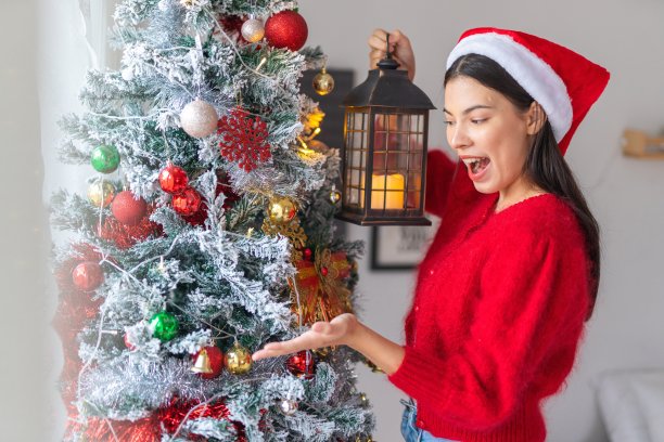 肖像照片的年轻美丽友好的亚洲女性女士与圣诞老人帽手持户外灯笼烛光旁边一个漂亮的装饰圣诞树图片下载