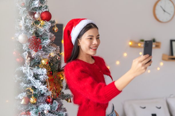 一位年轻美丽友好的亚洲女性女士的肖像照片，戴着可爱的圣诞老人帽，在她房间里装饰精美的圣诞树旁愉快地用她的智能手机自拍图片下载